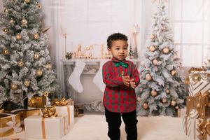 A young child in a red plaid shirt stands in front of Christmas decorations and wrapped presents.