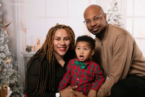 A family of three poses together for a Christmas portrait with decorated trees in the background.