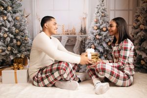 A couple in matching plaid pajamas shares a moment on the floor near a Christmas tree.