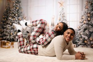 A couple in matching plaid pajamas plays and laughs on the floor by a Christmas tree.