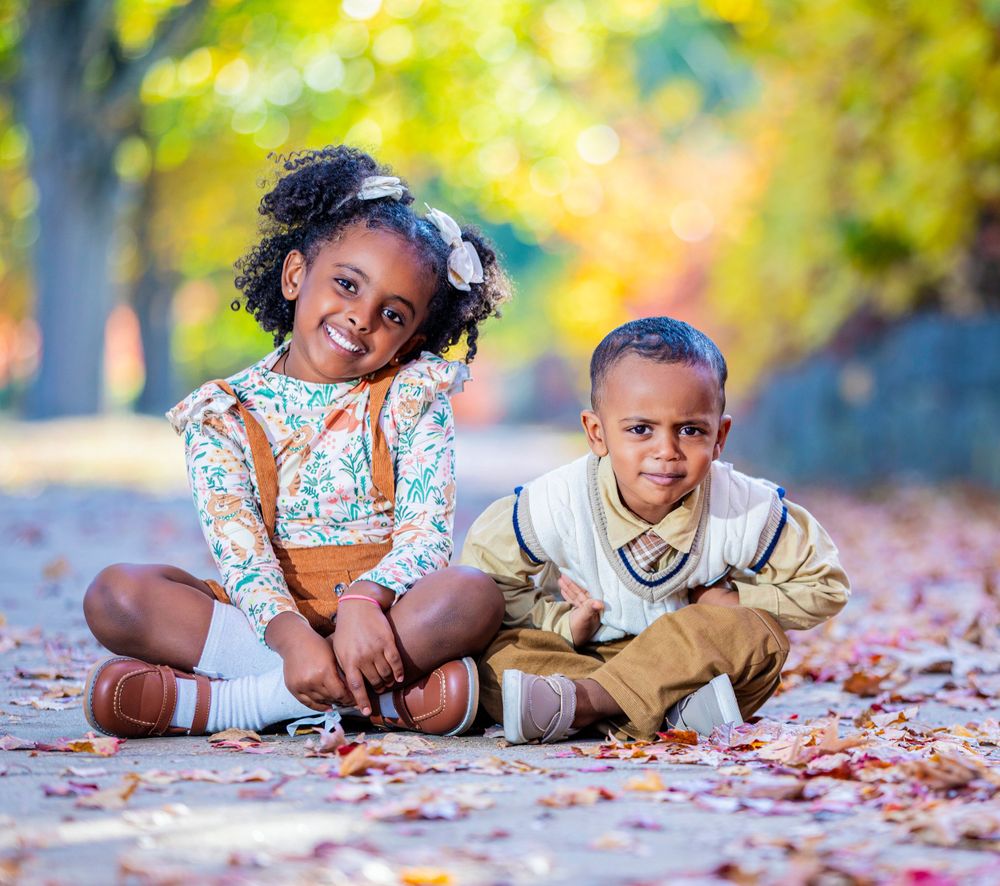 Two children sitting together on autumn leaves during outdoor photoshoot.