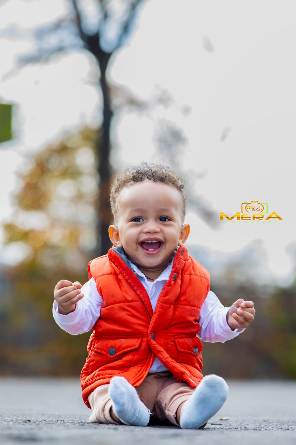 A young child wearing an orange vest and white shirt sits on the ground with a big smile.
