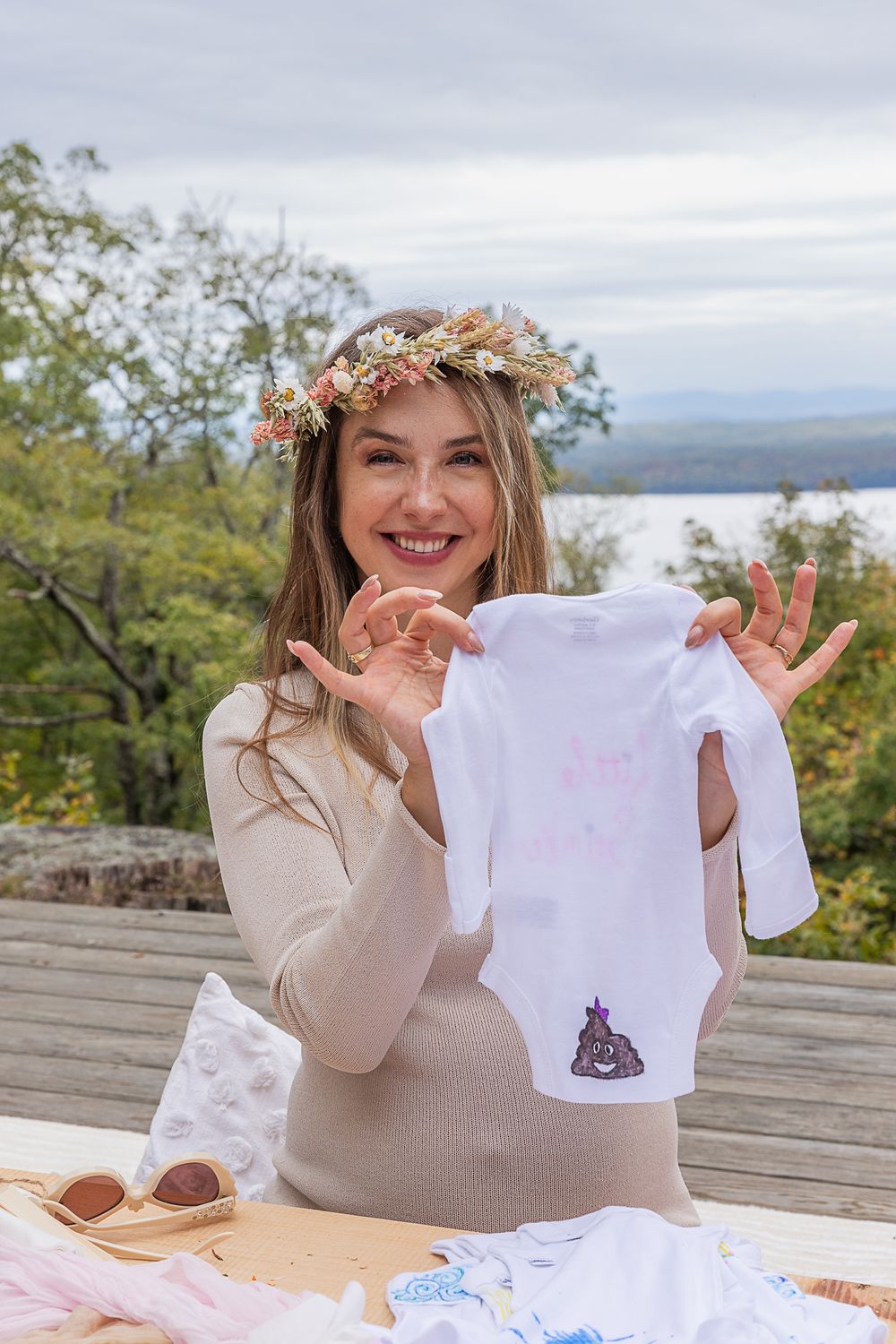 Person in cream sweater and flower crown holding up decorated baby onesie on outdoor deck.