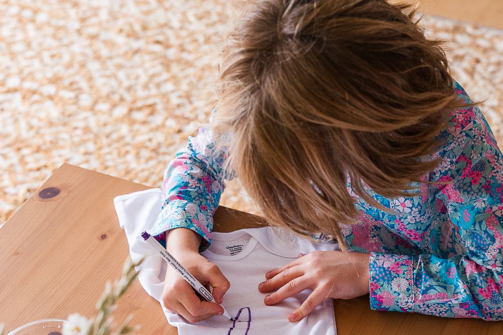 Child drawing on white fabric at wooden table during craft activity.