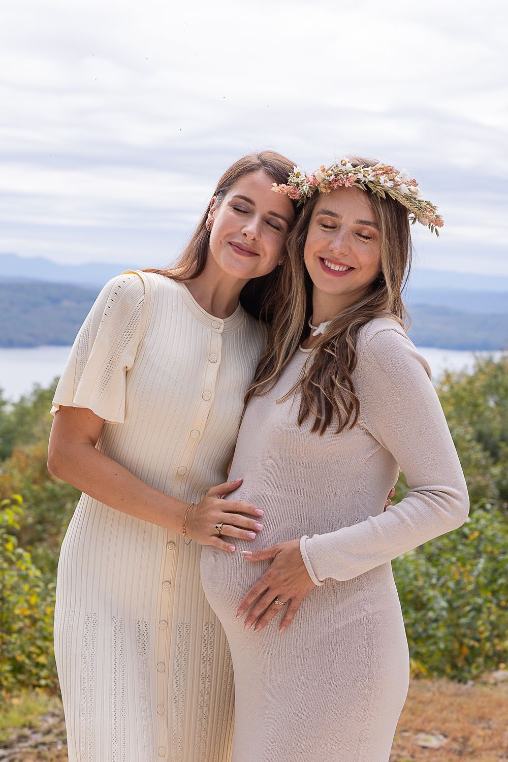 Two women in white dresses share tender moment against mountain backdrop wearing floral crowns.