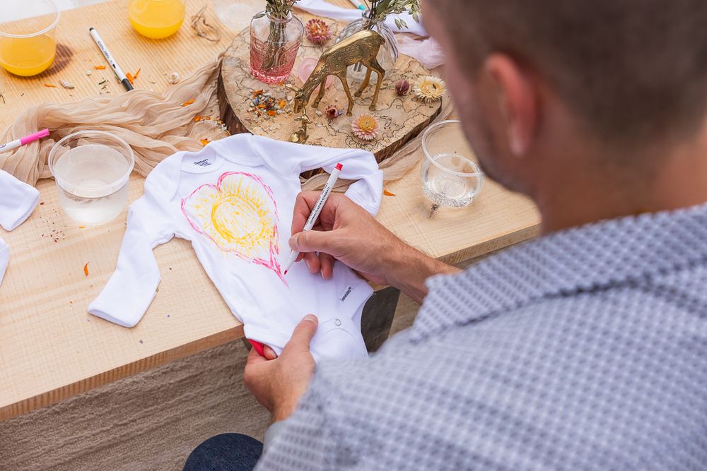 Person decorating a white baby onesie at a craft table with supplies.