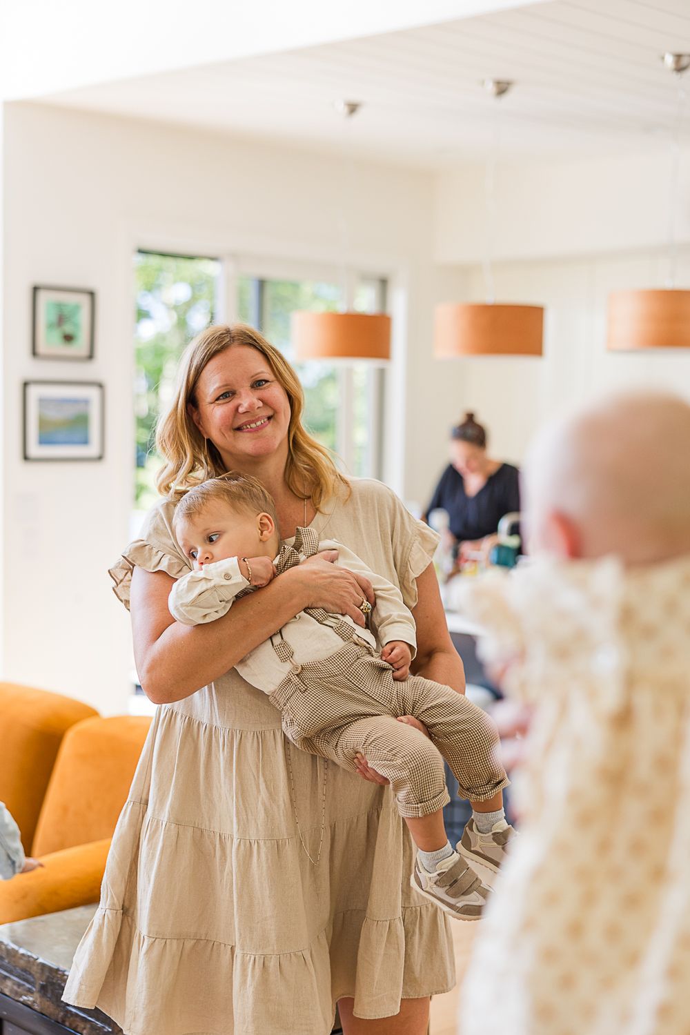 Woman in beige tiered dress holding an infant in a bright modern living room with orange pendant lights.
