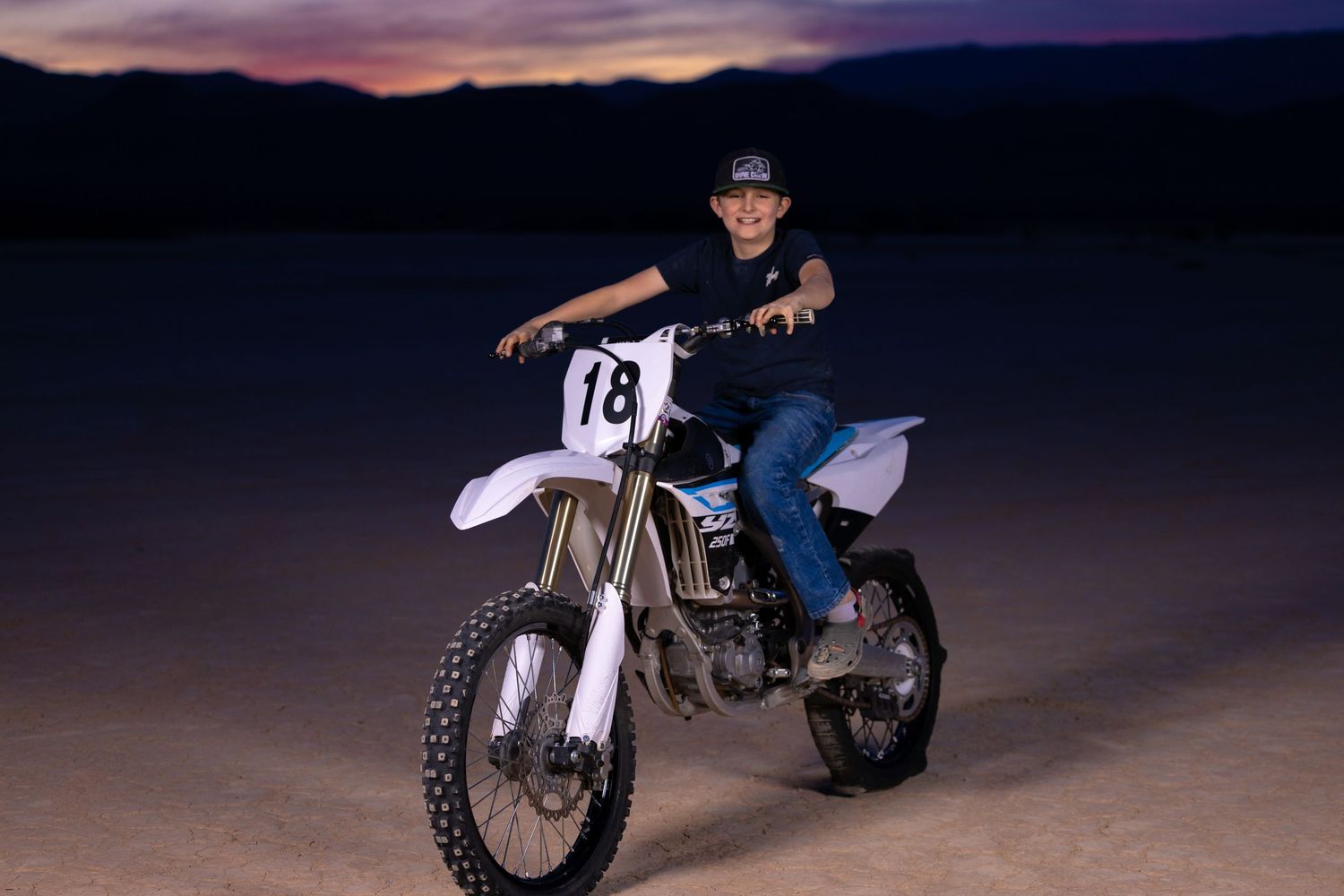 Dirt bike rider in black gear on a white numbered motorcycle at dusk against a purple sunset sky in desert terrain.