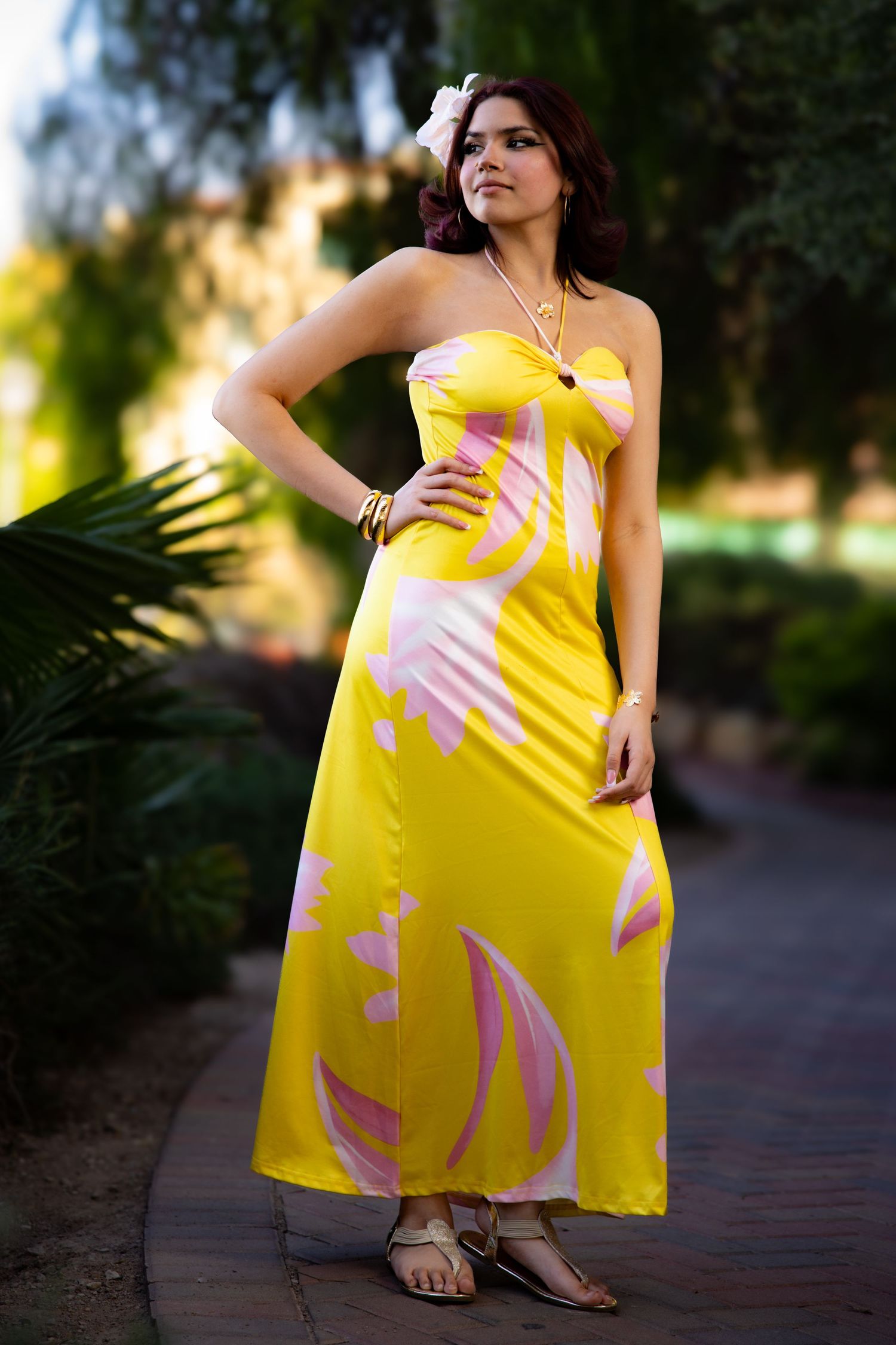 A woman poses outdoors in a bright yellow strapless maxi dress with pink abstract patterns and sandals.