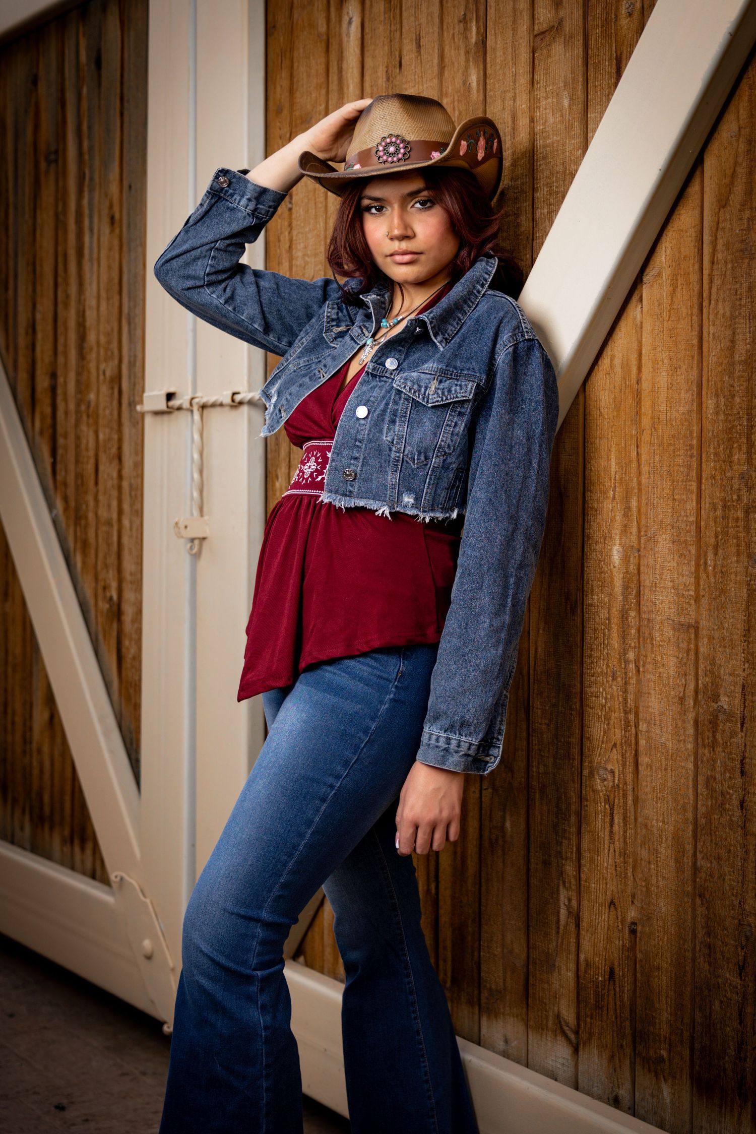 Western style outfit with cropped denim jacket, burgundy top, flared jeans and brown cowboy hat against wooden wall.