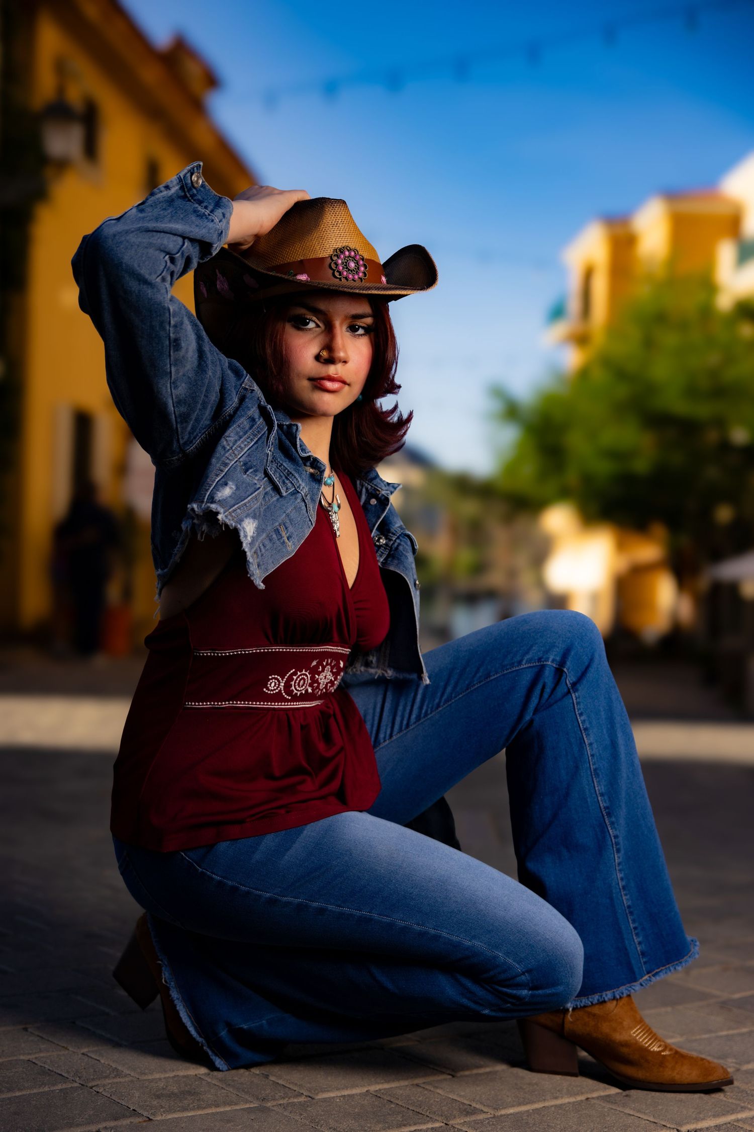 A model in western wear with cowboy hat and denim sits on cobblestones against yellow buildings.