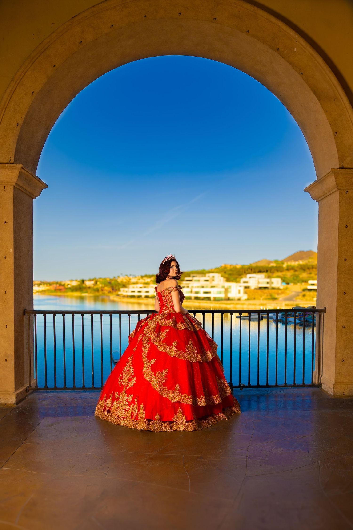A figure in an elegant red and gold quinceañera gown poses under a stone archway overlooking a waterfront view.