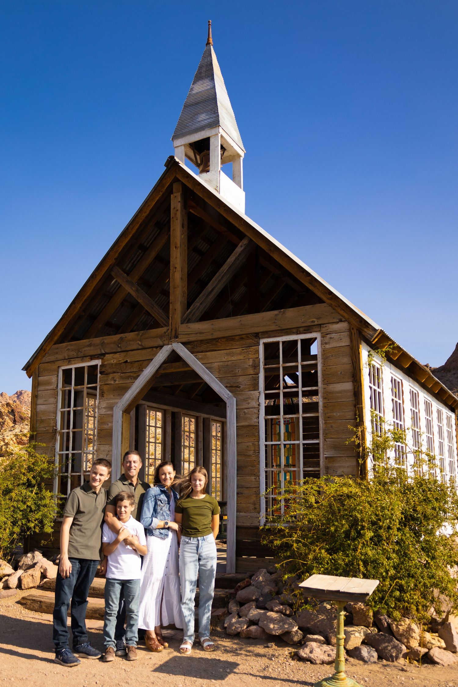 A group poses in front of a rustic wooden church building with white trim and a tall steeple against a blue sky.