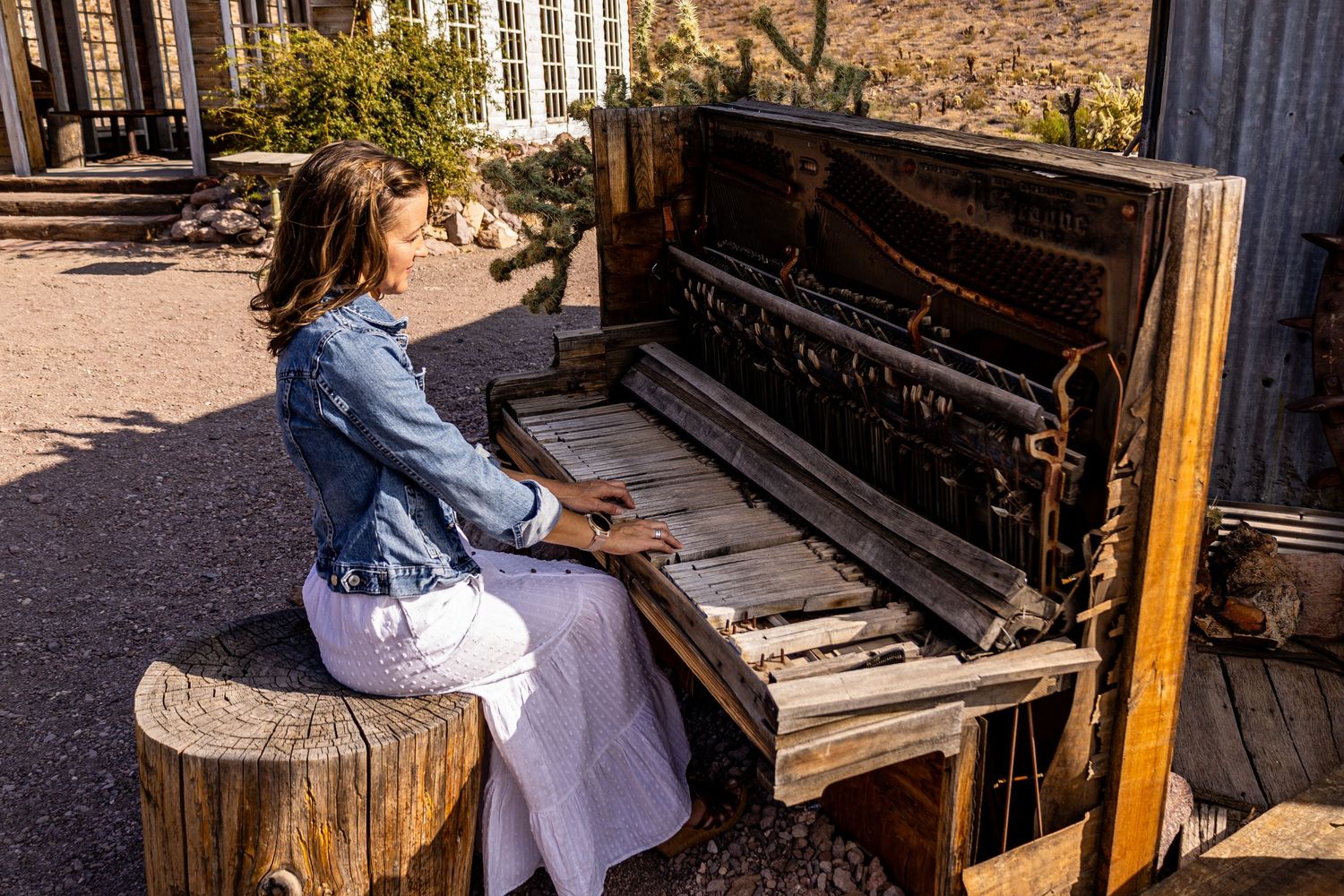 A person plays an old piano outdoors in a desert setting with warm sunlight casting shadows.