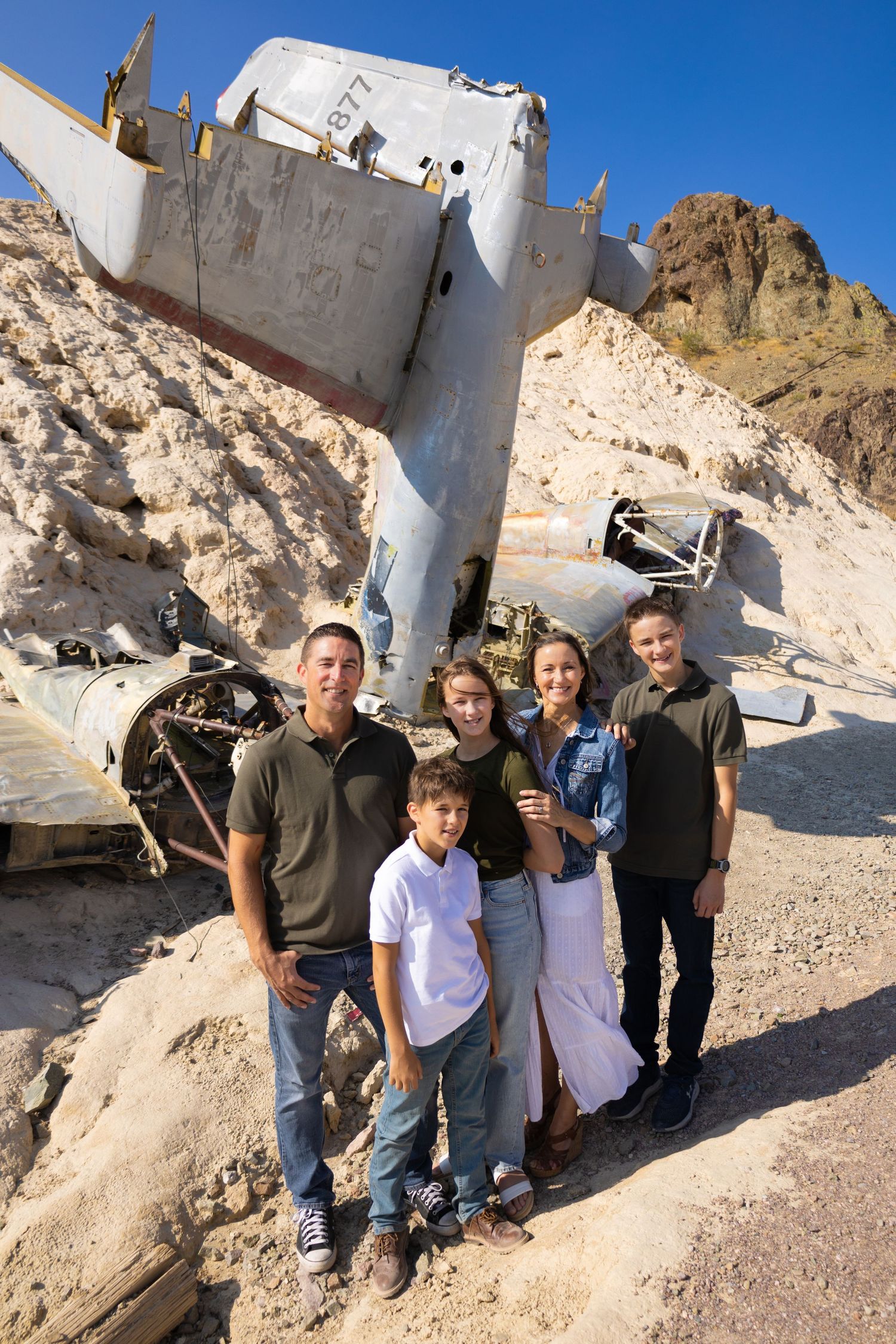 A group poses in front of a crashed airplane wreckage in a desert mountain setting.
