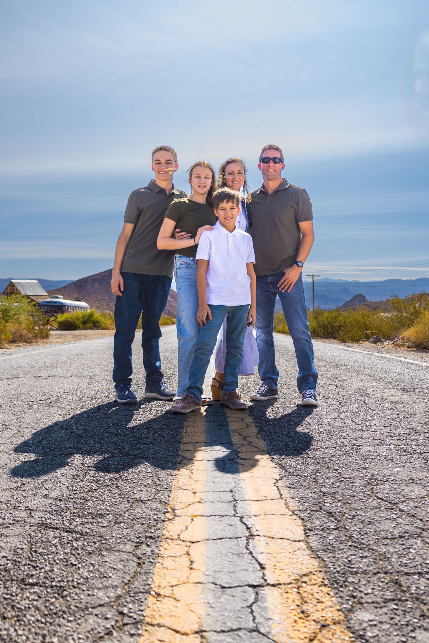 A group stands on a desert road with mountains and dramatic sky in the background.
