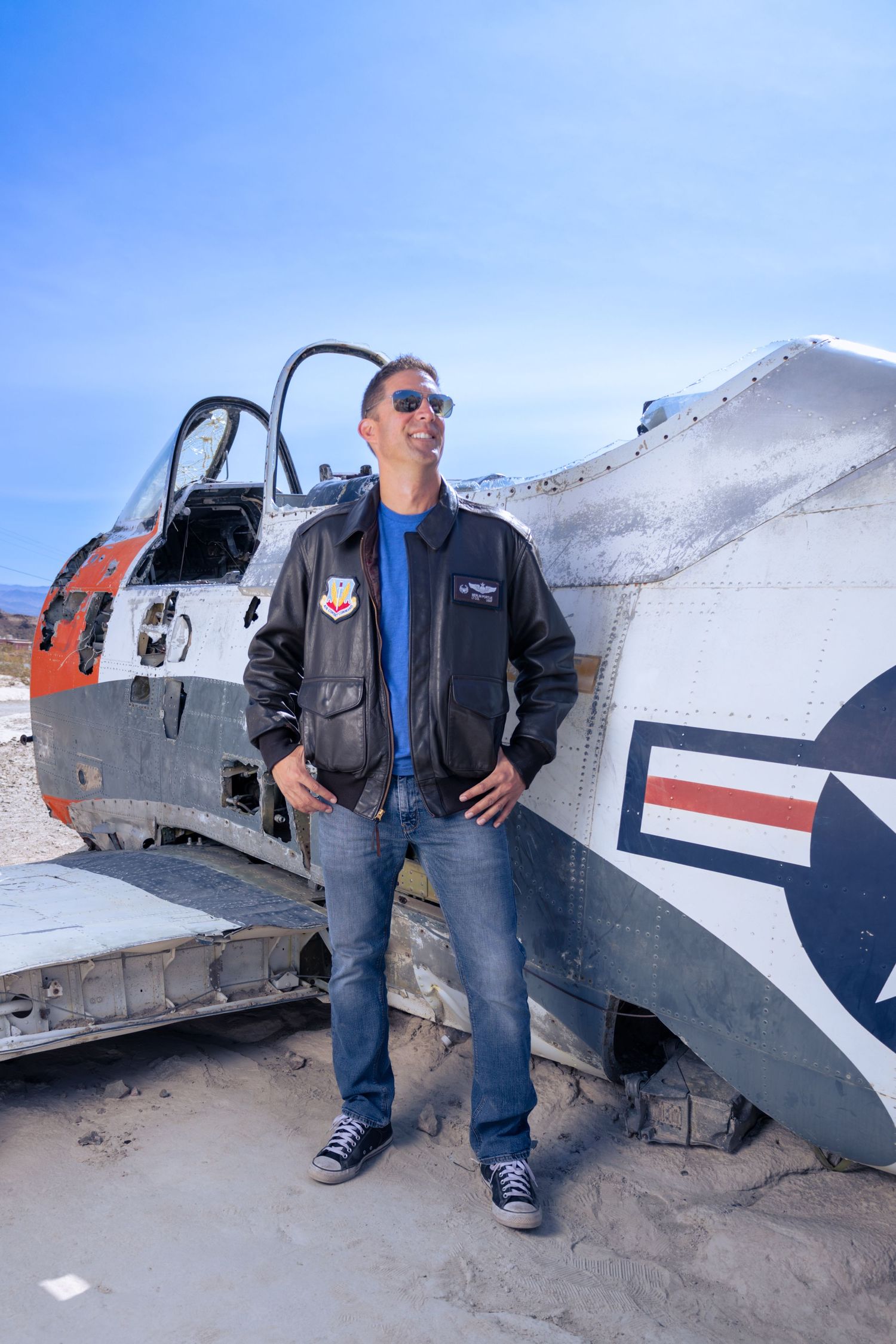 A person in a flight jacket poses next to a vintage military aircraft on a sunny day.