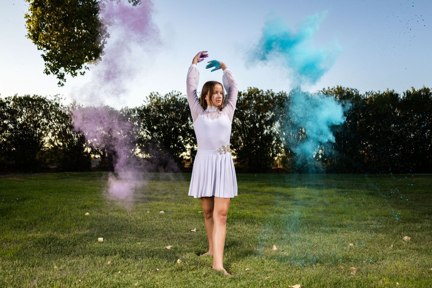 Dancer throws colorful powder while performing in white dress outdoors.