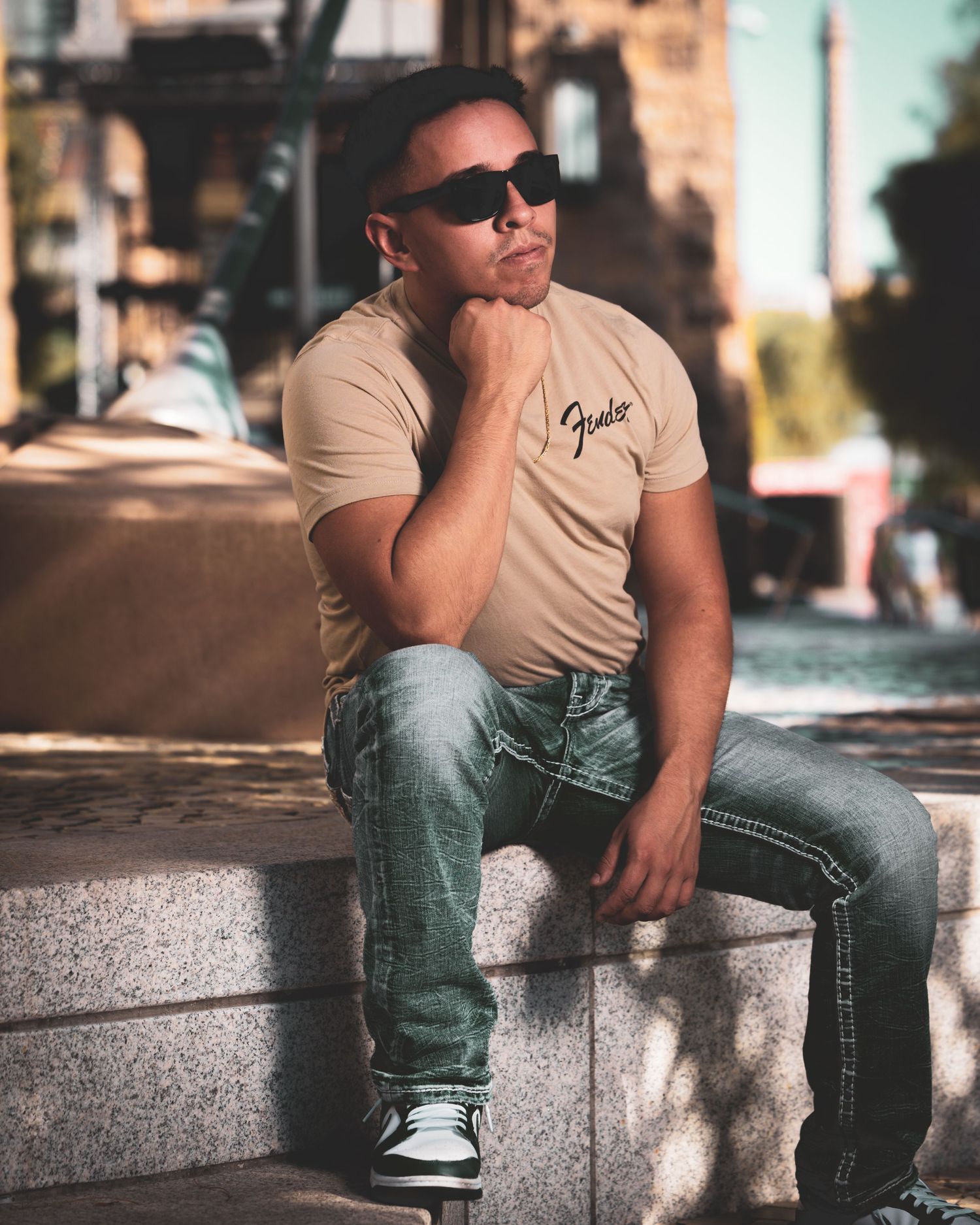 Urban portrait of person in beige t-shirt and jeans sitting on concrete wall wearing sunglasses.