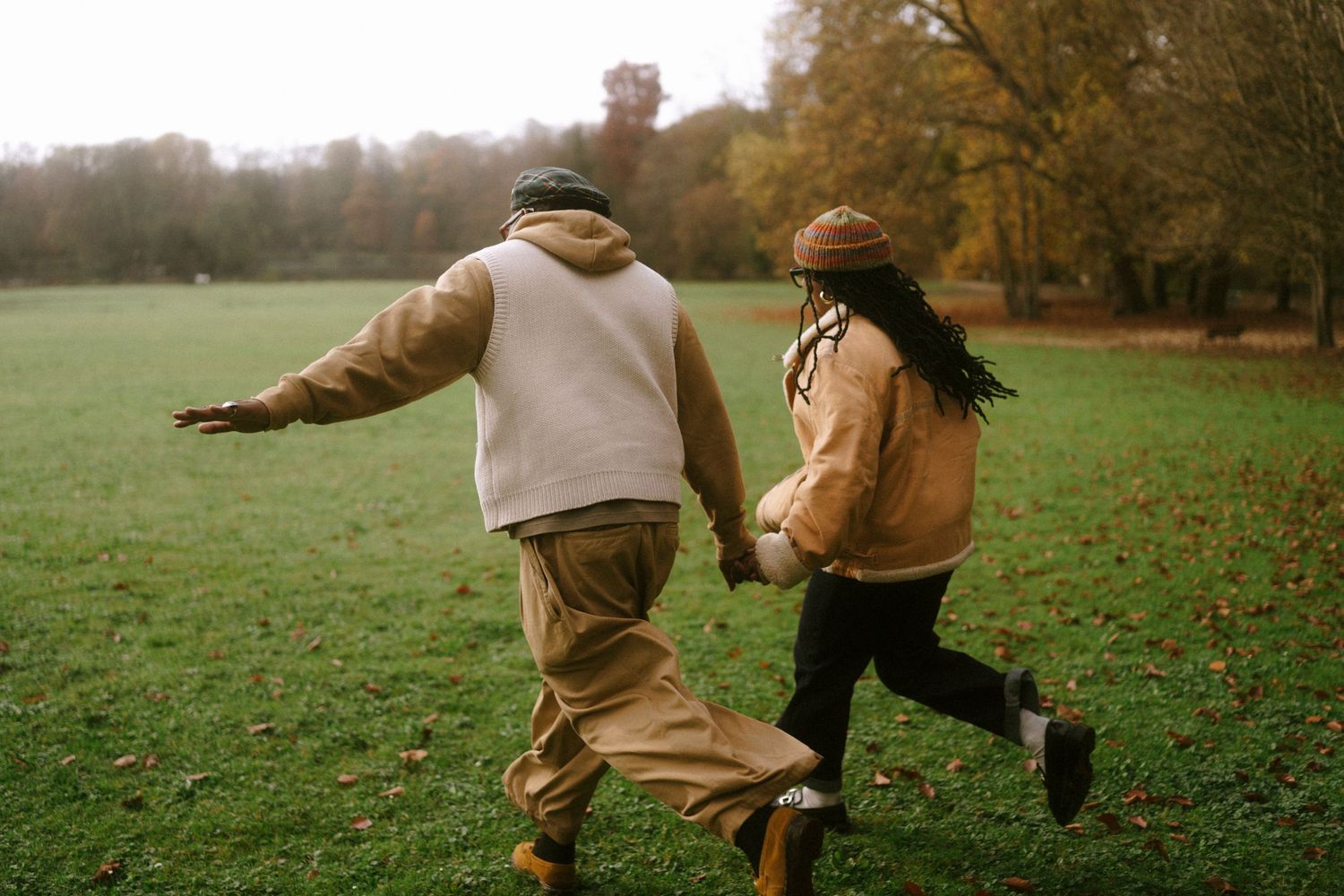 A couple runs playfully through an autumn field wearing matching brown jackets.