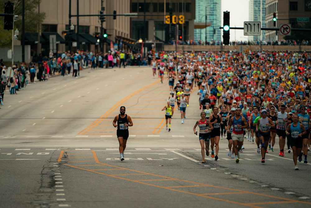 Marathon runners spread across a wide city street with tall buildings in the background.