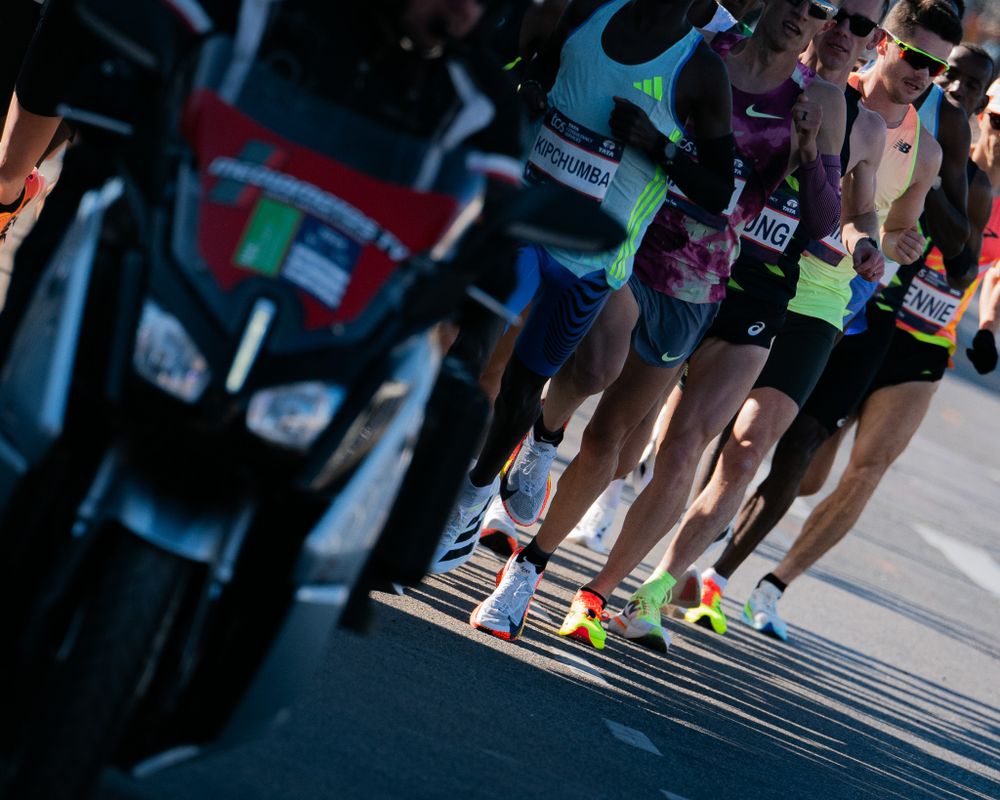 Marathon runners' legs in colorful running shoes casting shadows on the pavement during a race.