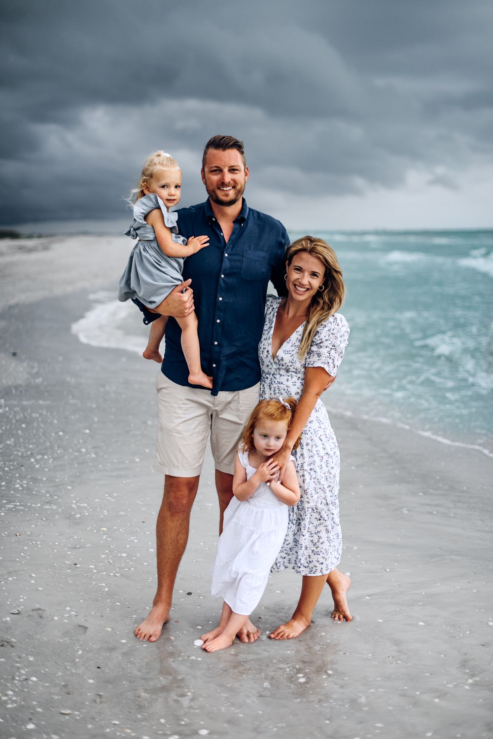Family of four poses together on stormy beach with turquoise waters in background.