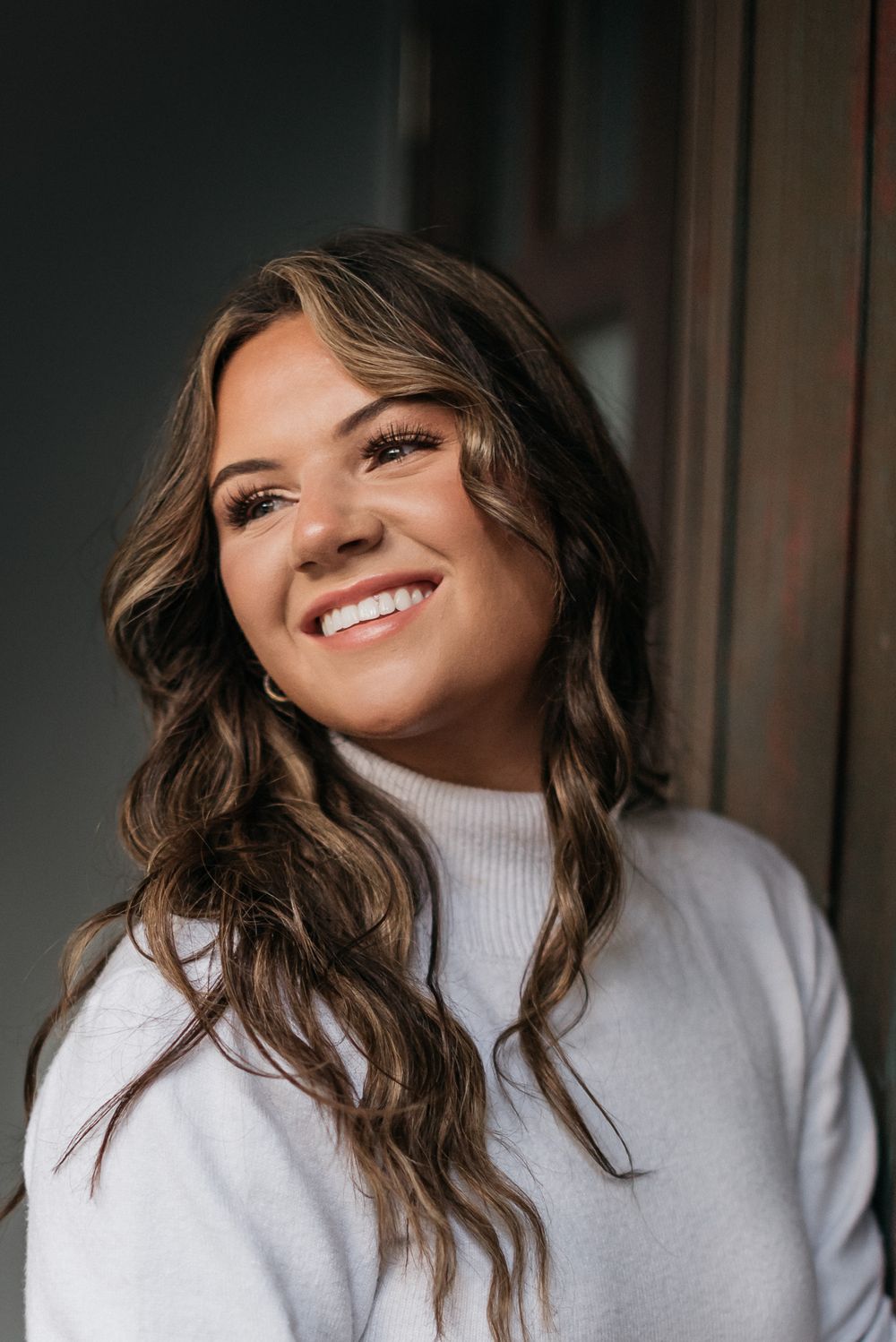 A portrait photo shows a bright smile and wavy brown hair against a dark indoor background.