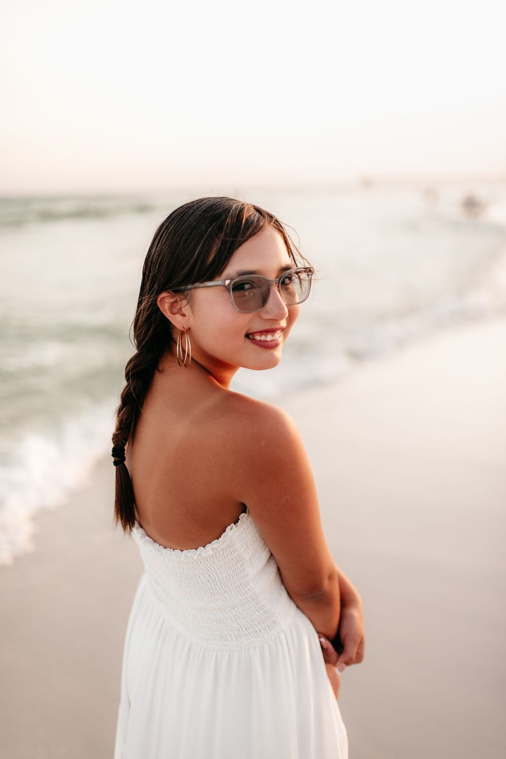 Young woman in white dress and sunglasses smiling at beach during golden hour.