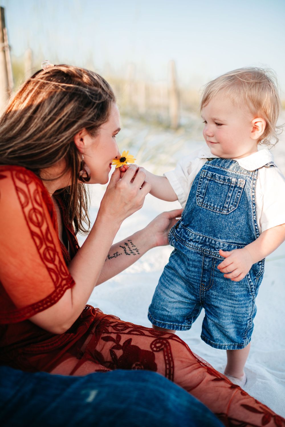 A sweet interaction between a toddler in overalls and woman in rust dress sharing a flower on the beach.