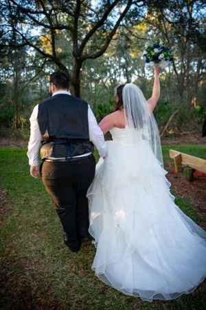 A newlywed couple walks outdoors, with the bride in a flowing white dress raising her arm in celebration.