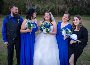 Wedding party wearing coordinated blue dresses and black attire pose together with bouquets outdoors.