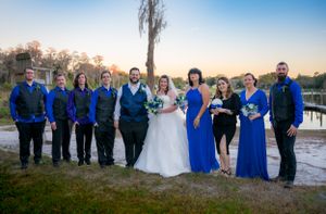 Wedding party in coordinating blue and black attire poses by a lake at sunset.