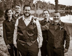 Wedding party members in vests and formal attire pose together by a waterfront.