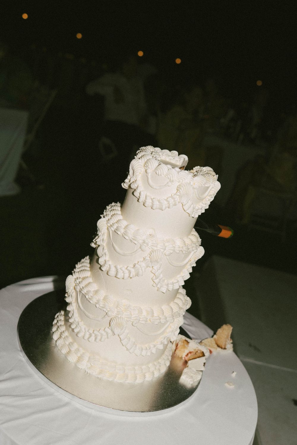 Elegant four-tier white wedding cake with ruffled frosting details on a silver stand, partially cut at a reception.
