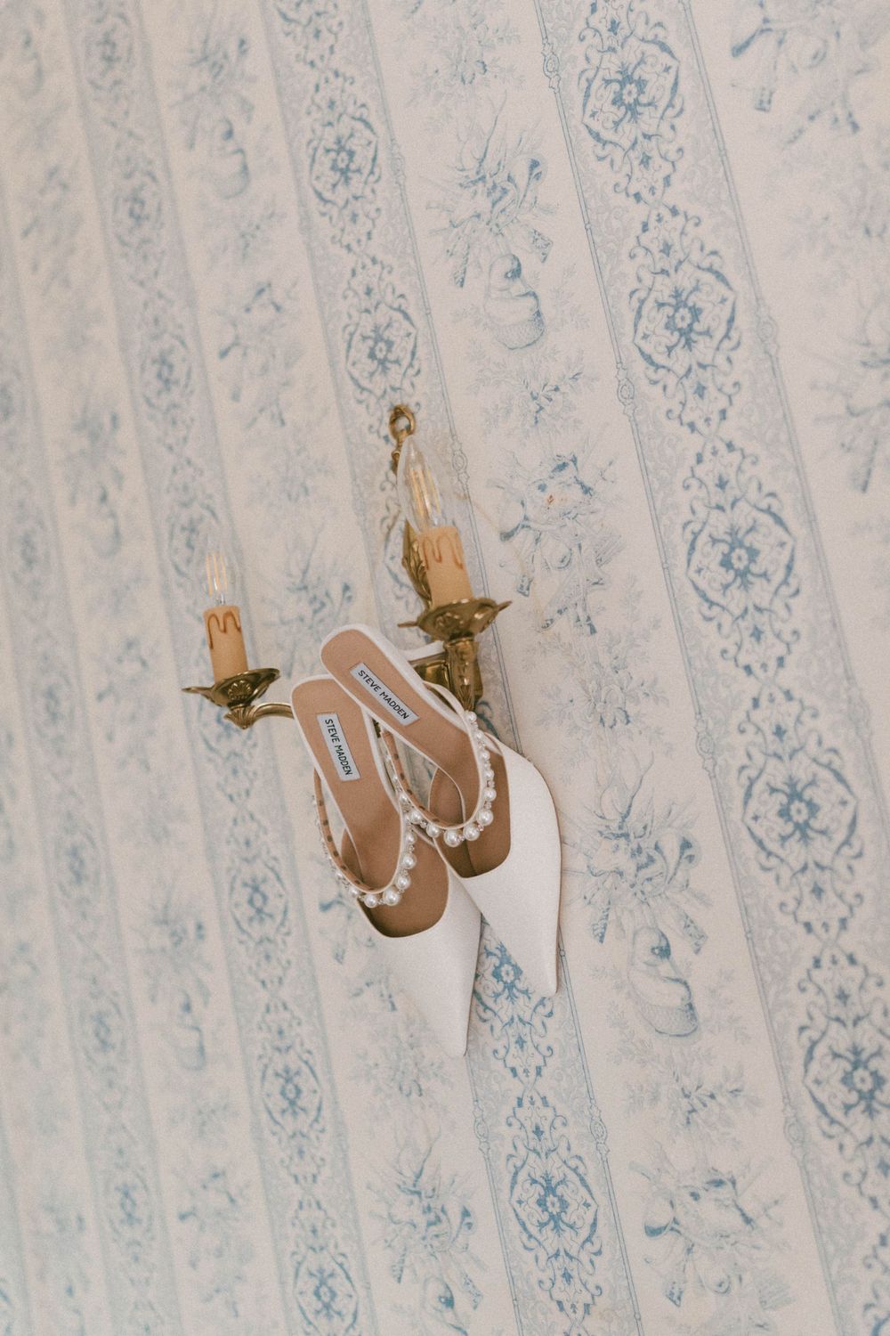 White bridal heels with pearl embellishments hanging on a gold wall sconce against blue toile wallpaper.