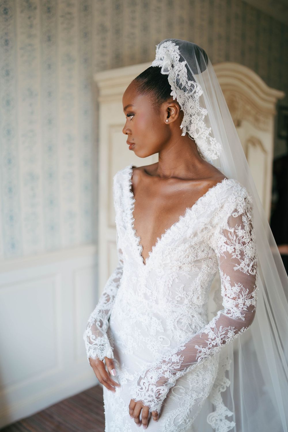 Elegant Black bride in lace long-sleeve wedding gown with floral veil cap, posing gracefully in a softly lit room.