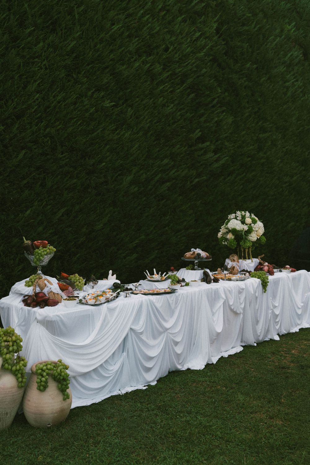 Elegant outdoor buffet table with white draped cloth, fresh fruit, flowers, and gourmet food against a lush green hedge backdrop.