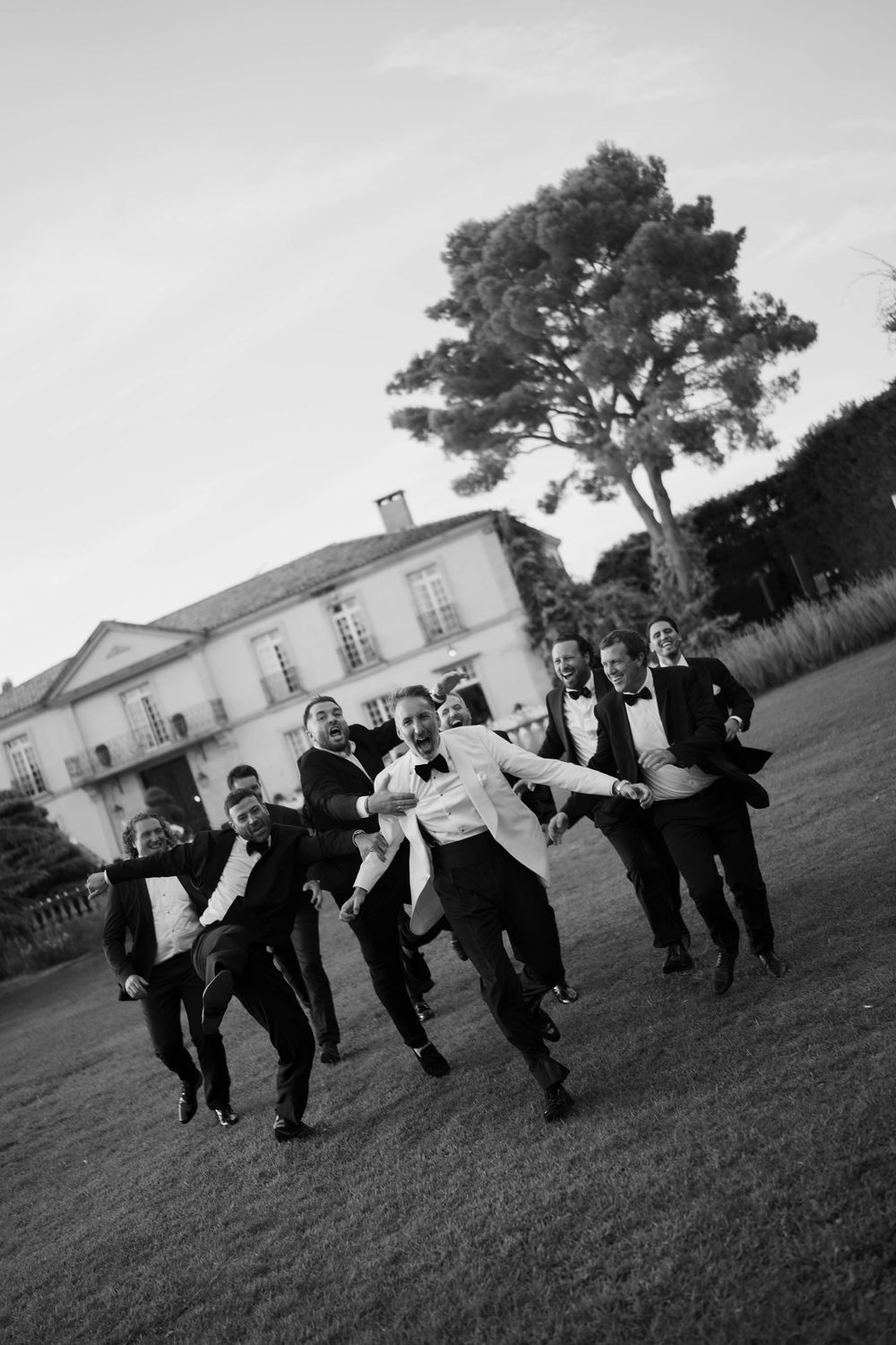 Black and white photo of groom and groomsmen in tuxedos joyfully running on lawn outside elegant estate.