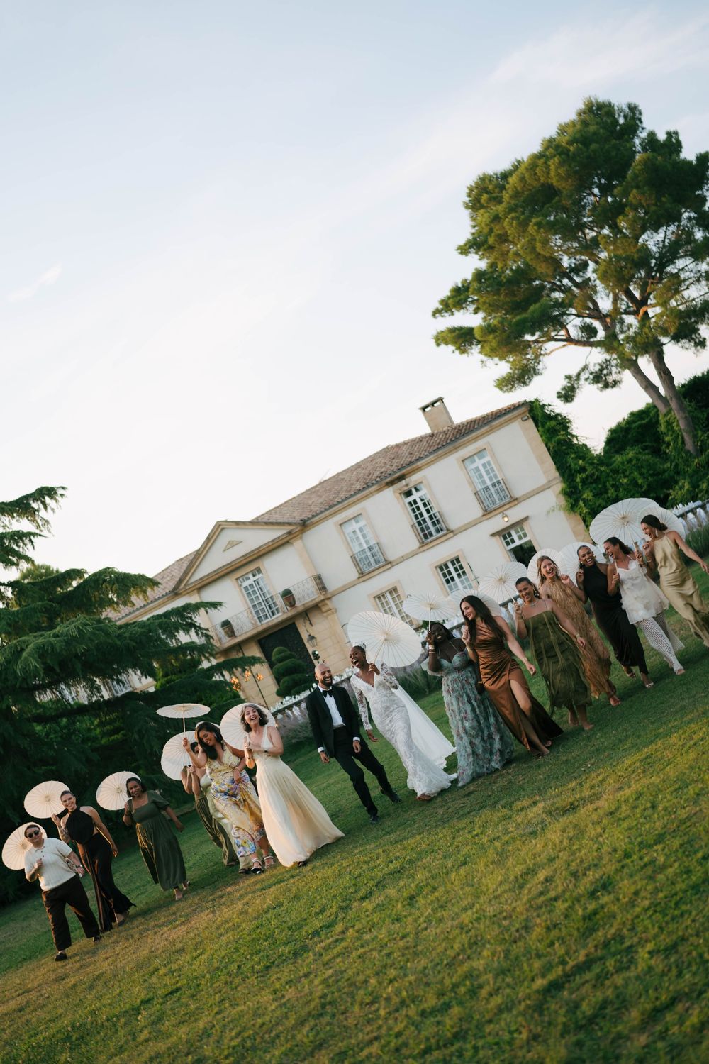 Wedding party celebrating on lawn before elegant French chateau at dusk, bridesmaids in colorful gowns holding parasols.