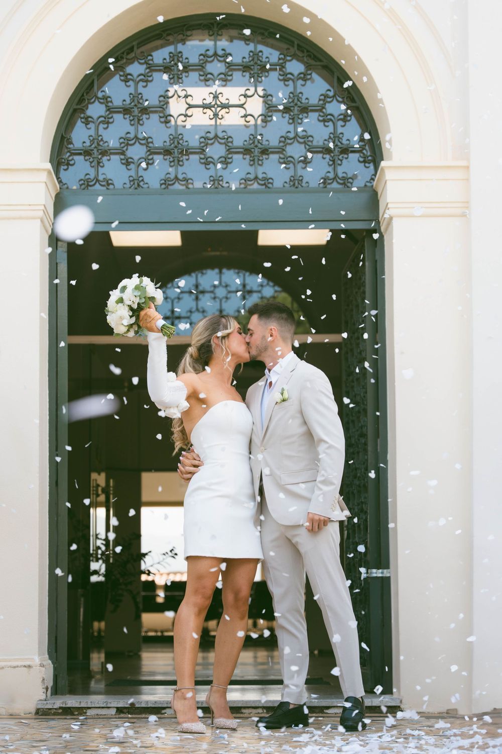 A couple shares a celebratory kiss in front of an ornate doorway as white confetti falls around them.