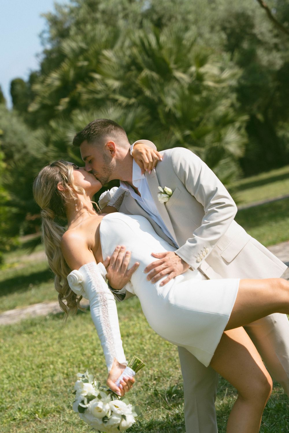 A romantic outdoor wedding scene with a couple sharing a dip kiss while holding a white floral bouquet.