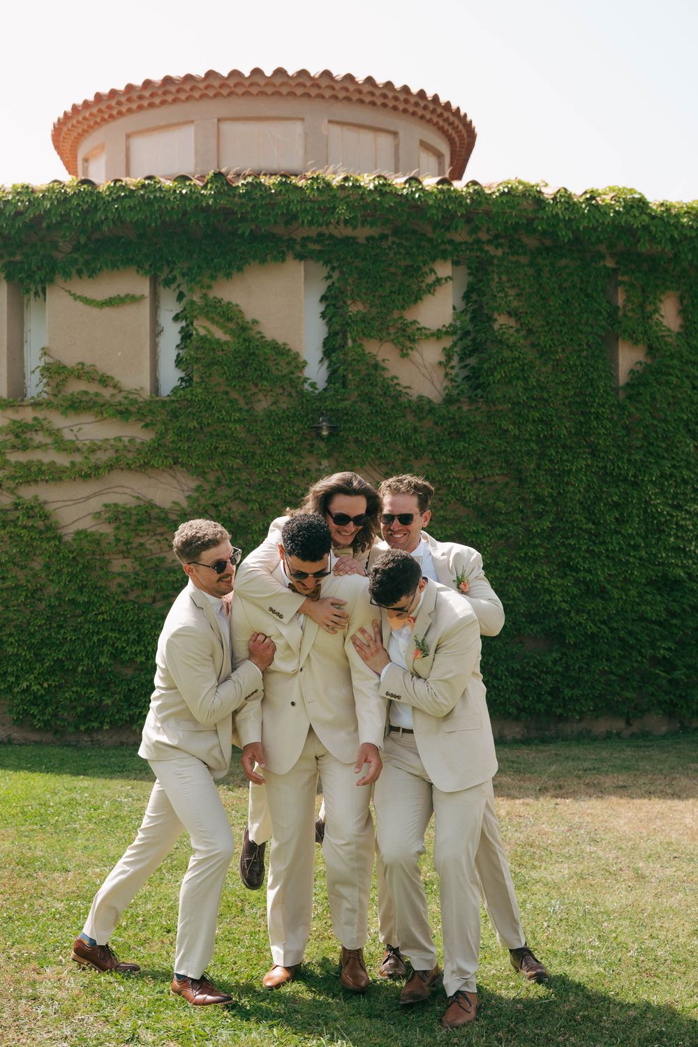 A group in matching cream-colored suits huddle together on a lawn in front of an ivy-covered building.