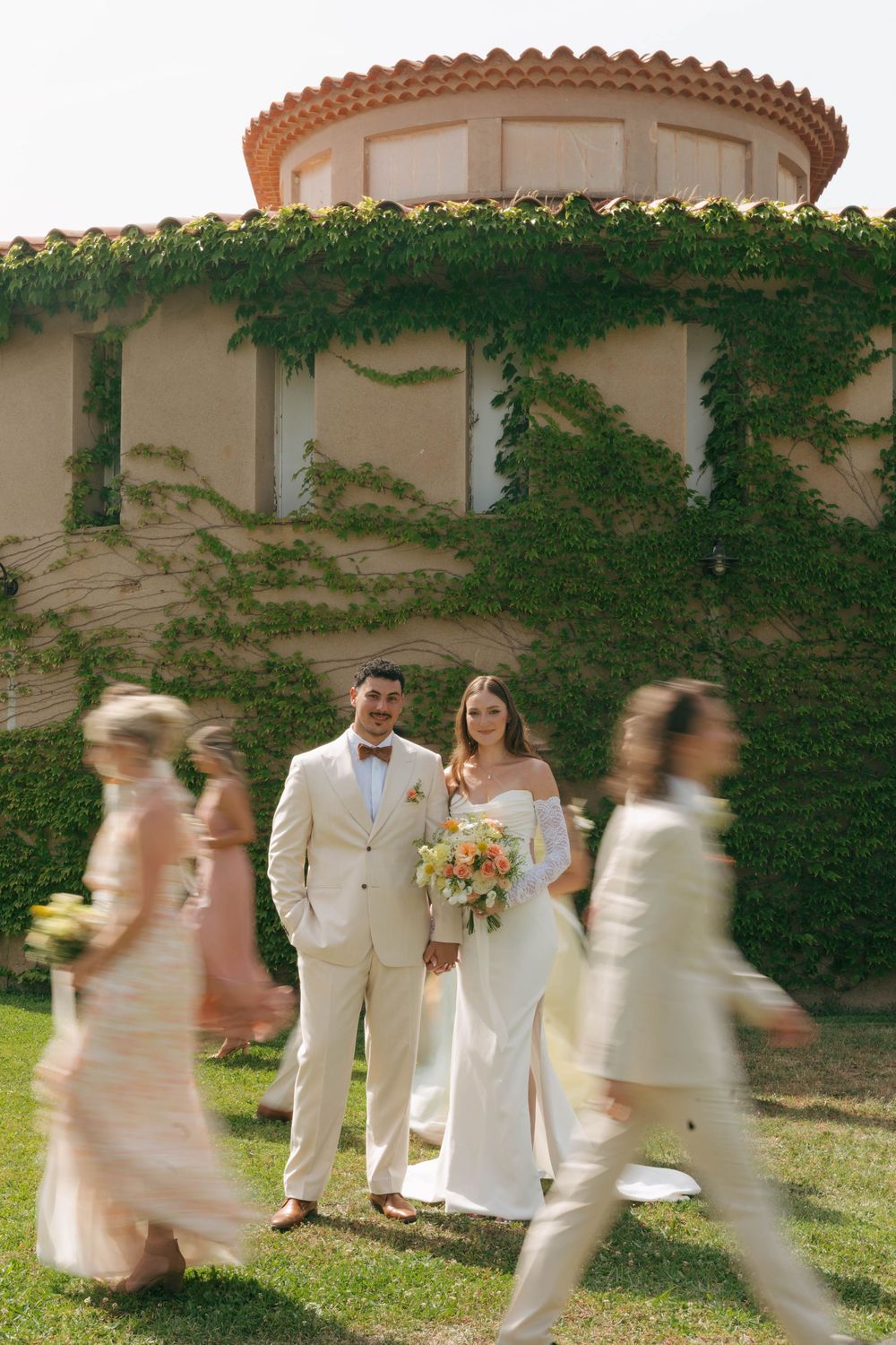 Wedding couple standing in front of an ivy-covered villa with a terracotta dome as guests walk past on a sunny day.