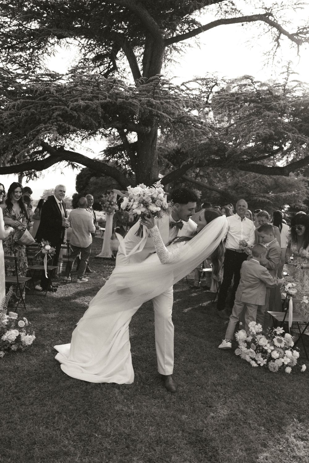 A couple performs a romantic dip during their first dance at an outdoor garden wedding under a large tree.