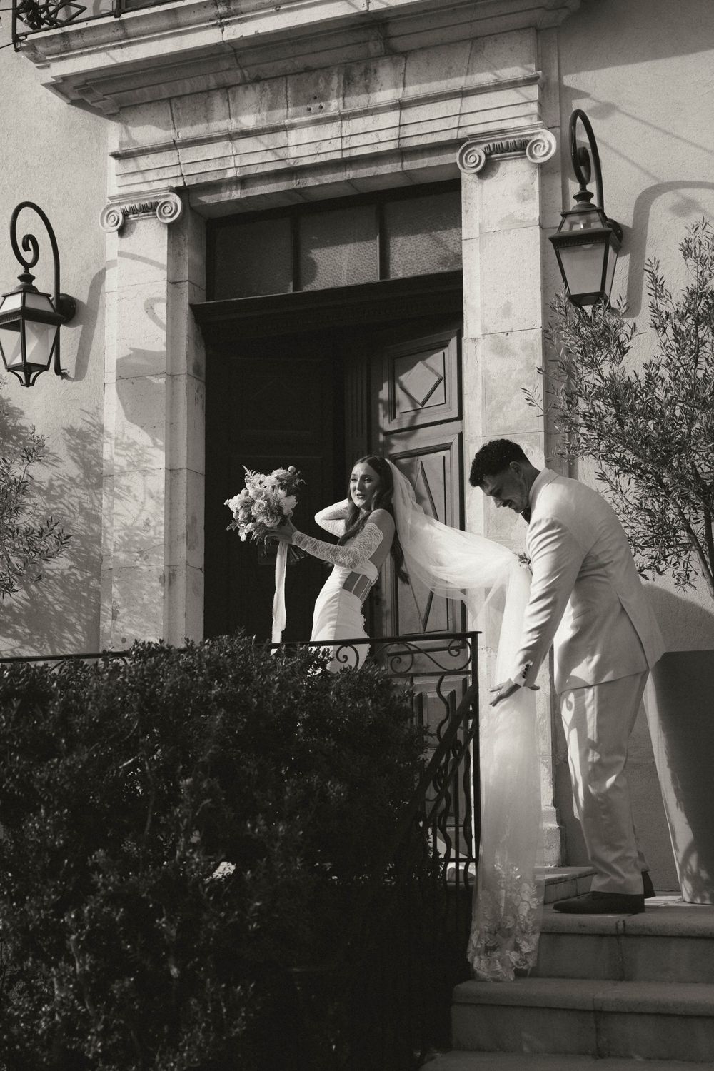 A couple in formal attire stands on the steps of an elegant entrance with lanterns and decorative stonework.