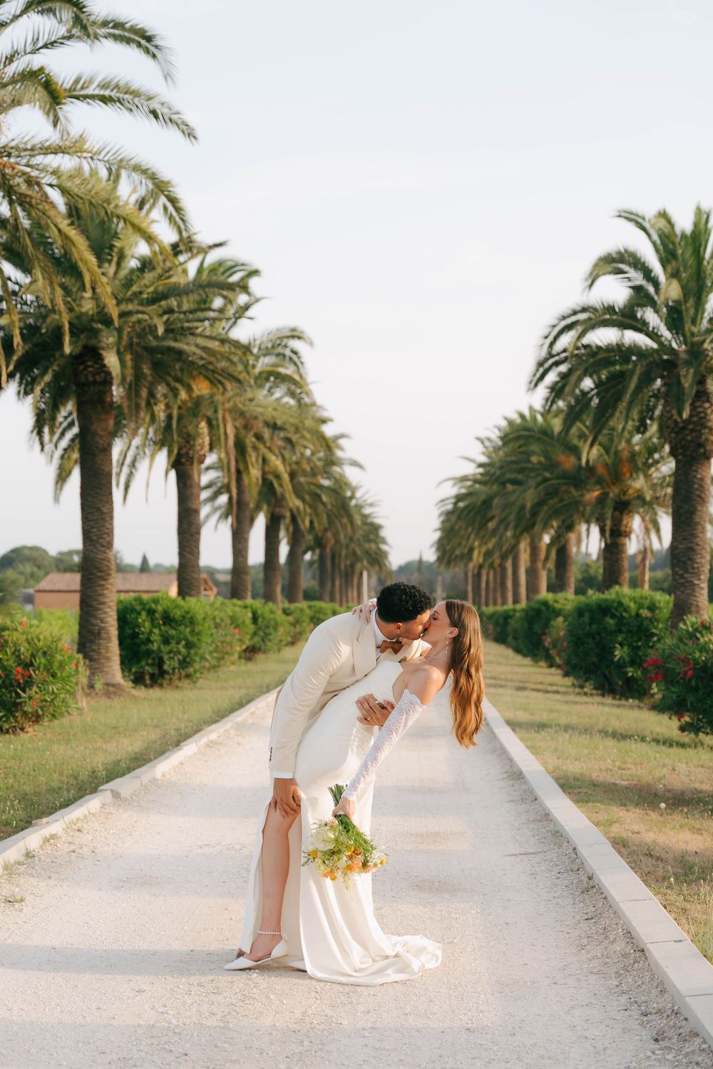 Romantic couple in wedding attire sharing a kiss on a palm tree-lined pathway at sunset.