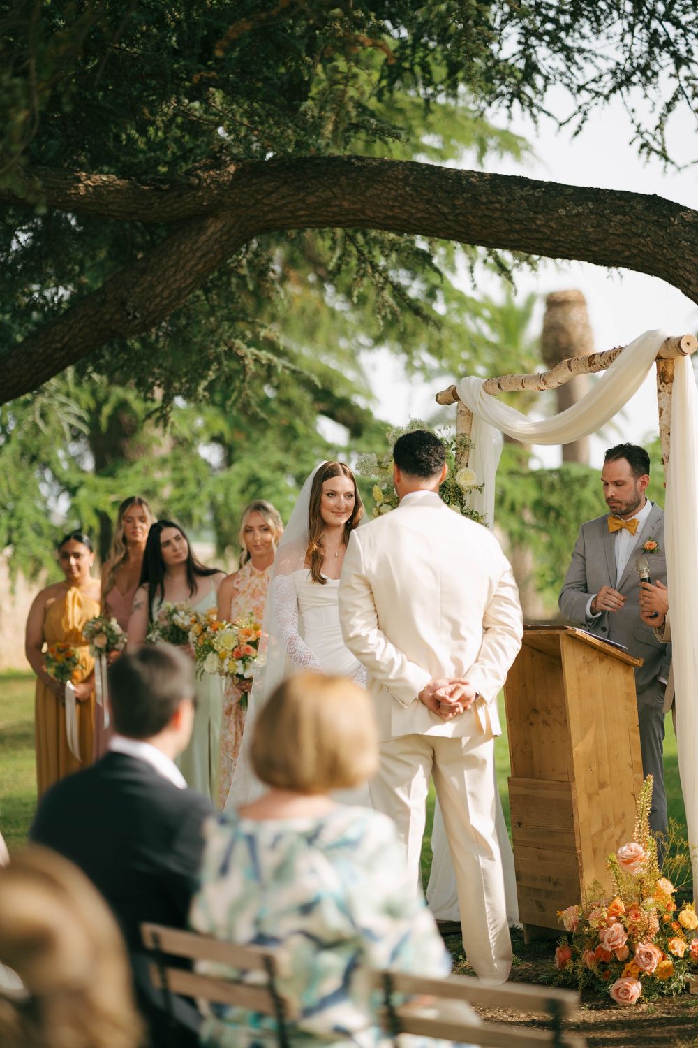 Wedding ceremony takes place under rustic wooden beams draped with white fabric and floral arrangements at sunset.