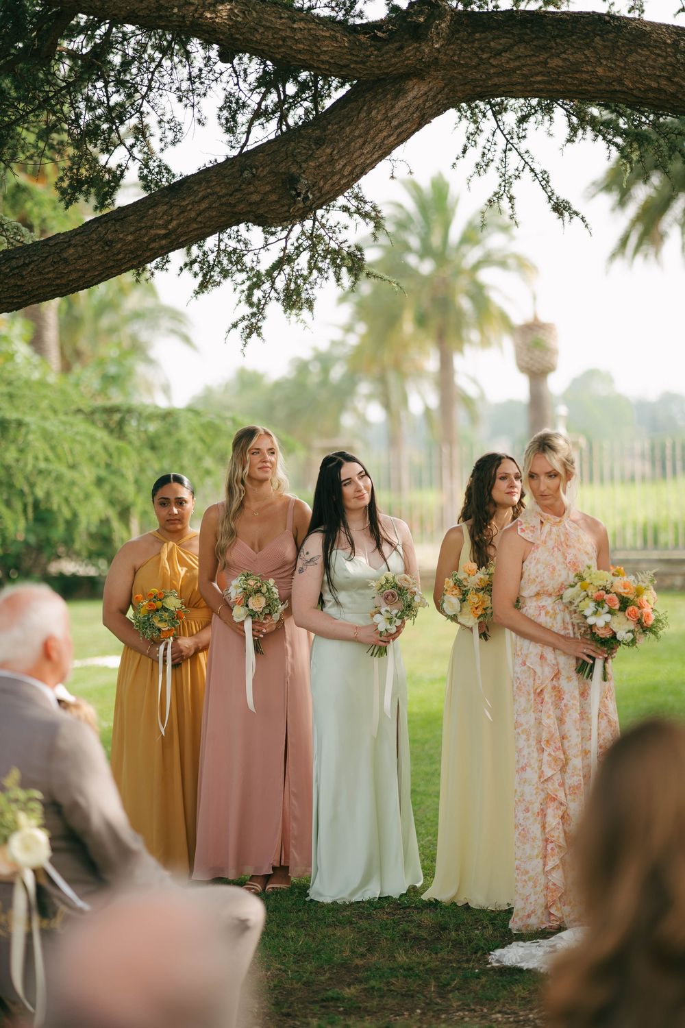 Bridal party wearing mixed pastel dresses stands together under a large oak tree at sunset in a tropical setting.