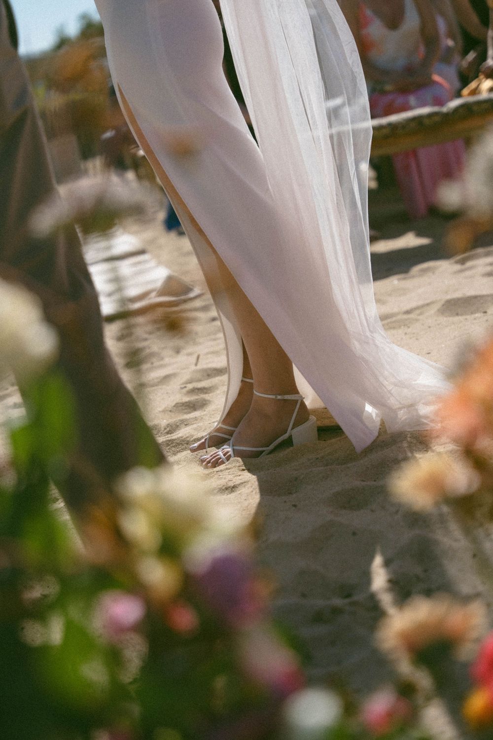 Close-up of bride's feet in white shoes on sandy beach with flowing white dress and blurred flowers in foreground.